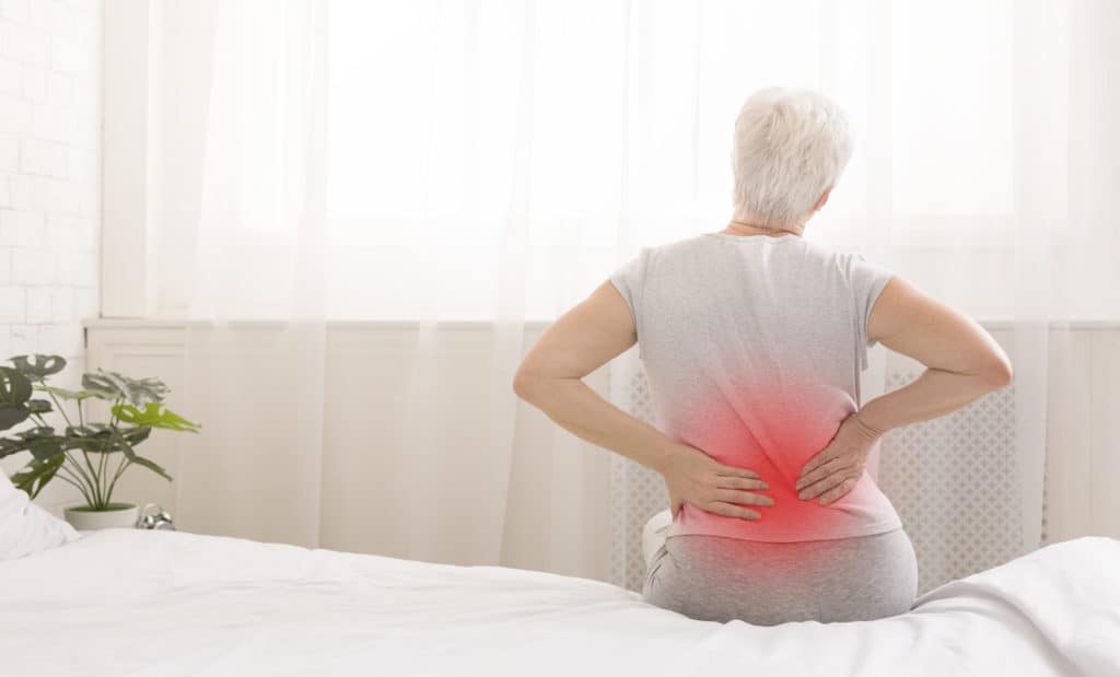 Senior woman suffering from backache in morning sitting on bed, red sore zone, panorama with free space
