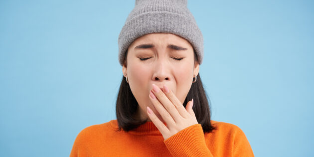 a woman in an orange sweater and grey hat with her hand over her mouth wondering, why am i alsys tired?